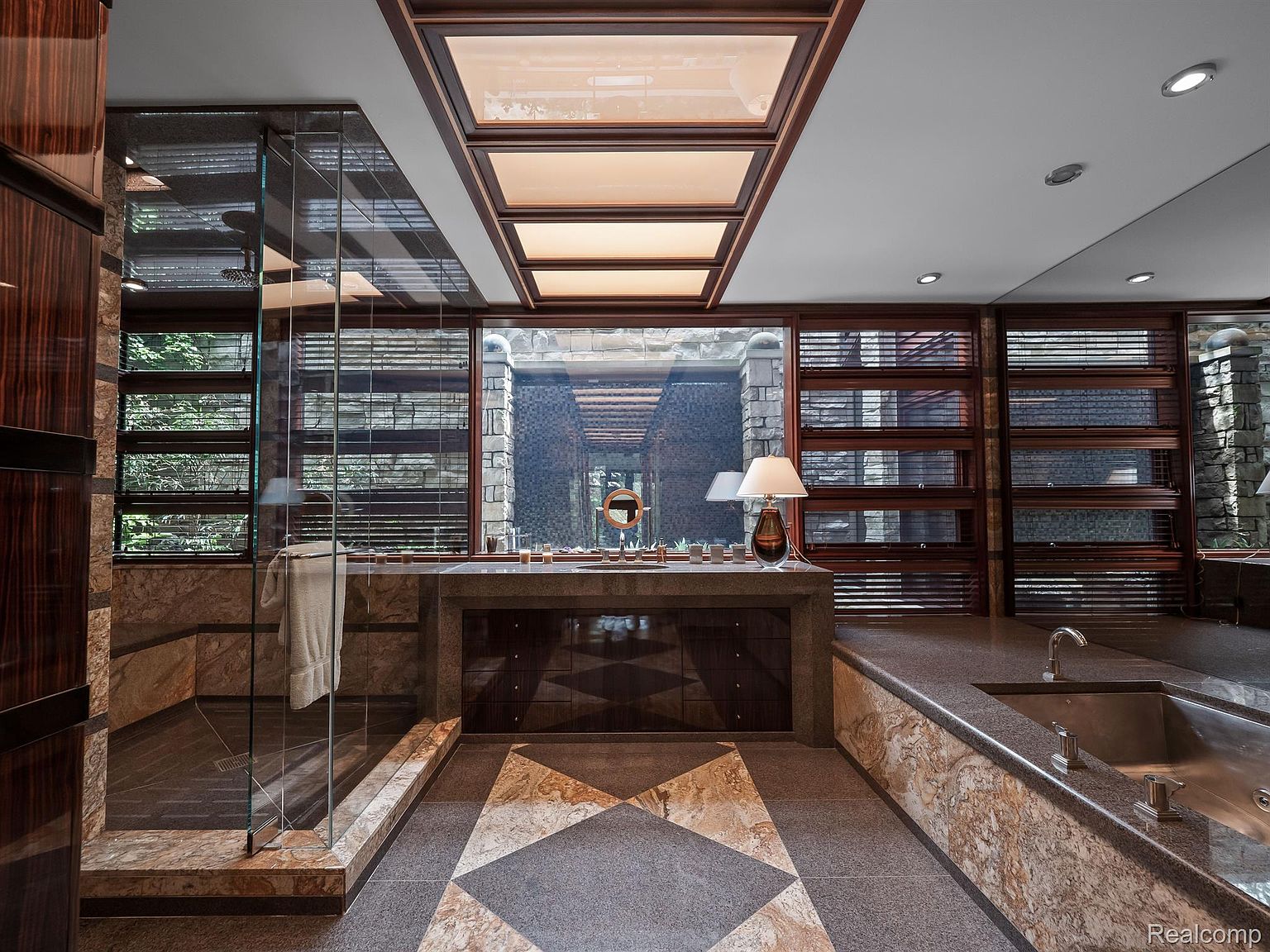 This is a luxurious primary bathroom featuring a walk-in shower with glass enclosure, a large vanity with a stone countertop, and a built-in bathtub. The room is illuminated by natural light from a skylight and windows with wooden blinds, creating a spa-like atmosphere. The flooring is a patterned tile, and the overall design is sophisticated and modern.