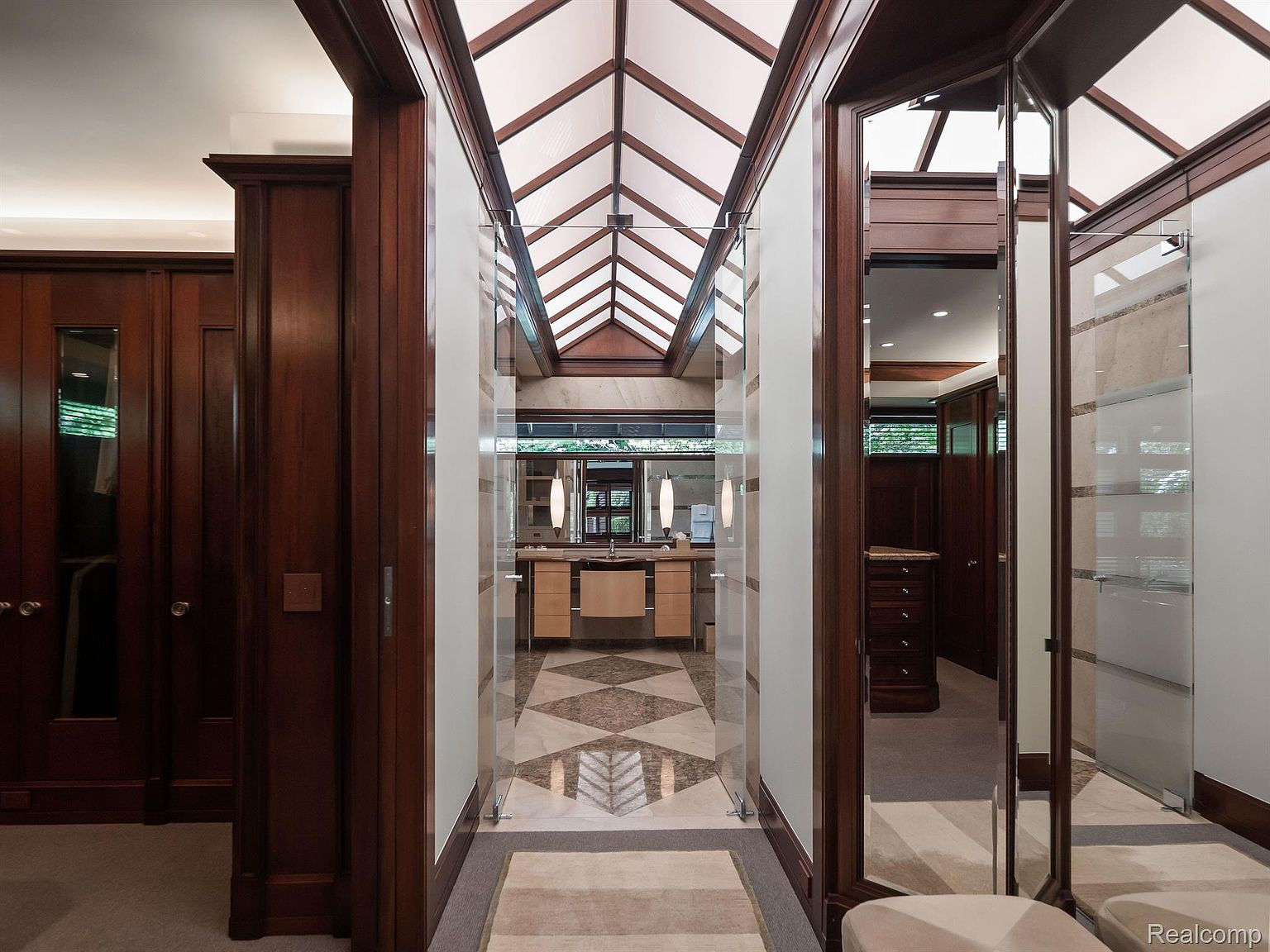 This is a luxurious primary bathroom featuring a unique skylight design that floods the space with natural light. The bathroom includes a vanity area with modern fixtures, a patterned tile floor, and glass-enclosed shower or toilet areas on either side. The dark wood trim and cabinetry add a touch of elegance and sophistication.