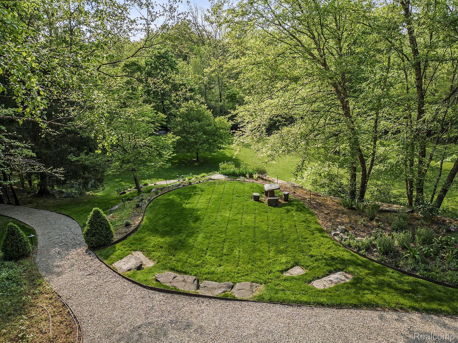 This is an aerial view of a meticulously maintained yard and garden. The focal point is a lush green lawn bordered by a gravel path and mature trees. A stone seating area adds a touch of rustic charm, creating an inviting outdoor space perfect for relaxation and entertaining.