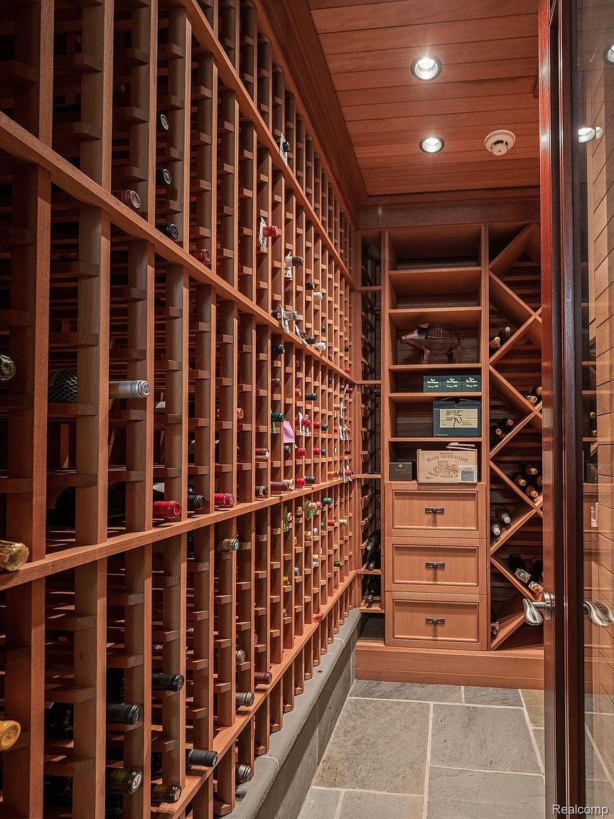 This is an interior shot of a well-organized wine cellar. The cellar features extensive wooden racking systems filled with wine bottles, a stone floor, and built-in cabinetry with drawers and shelves. The warm wood tones and strategic lighting create a sophisticated and inviting atmosphere, perfect for showcasing a wine collection.