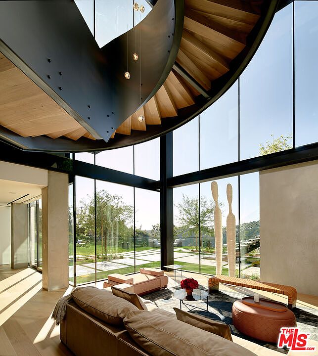 This is a stunning interior shot of a modern living room featuring floor-to-ceiling windows that offer expansive views of the exterior landscape. A unique spiral staircase adds an architectural focal point, while contemporary furniture and decor create a sophisticated and inviting atmosphere. The room is bathed in natural light, enhancing the sense of openness and luxury.