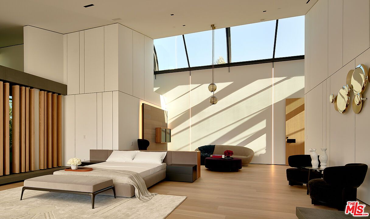 This is a spacious primary bedroom featuring a modern design with clean lines and a minimalist aesthetic. The room includes a large bed, a seating area with a curved sofa and armchairs, and unique architectural details such as a high ceiling with a skylight and decorative wall mirrors. The hardwood flooring and neutral color palette create a serene and luxurious atmosphere.