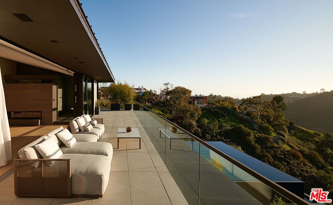This image showcases a luxurious outdoor patio or balcony area, featuring modern furniture such as plush sofas and a sleek coffee table. The space is defined by a glass railing, offering unobstructed views of the surrounding hillside landscape and distant houses. The design emphasizes a seamless indoor-outdoor transition, perfect for relaxation and entertainment.