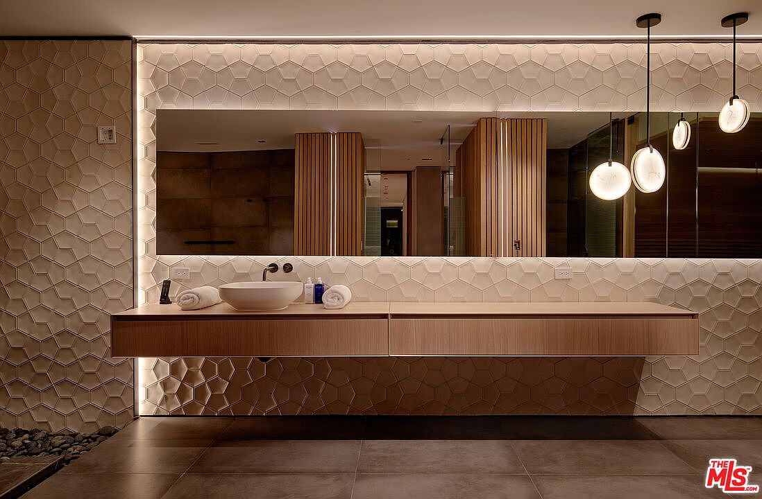 This is a modern primary bathroom featuring a floating vanity with a vessel sink and sleek faucet. The wall is adorned with textured, geometric tiles, and a large mirror with integrated lighting enhances the space. Pendant lights hang above the vanity, adding a touch of elegance.