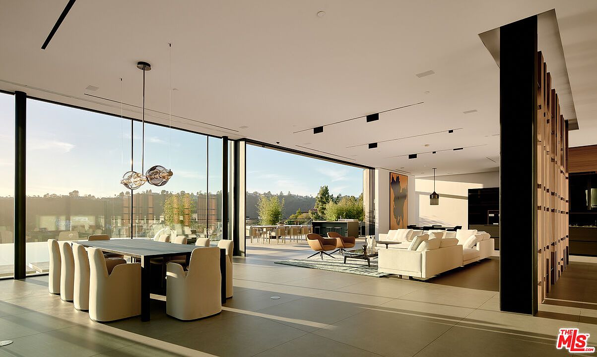 This is a spacious and modern living room featuring floor-to-ceiling windows that offer panoramic views of the outdoors. The room includes a dining area with a large table and chairs, a comfortable seating area with sofas and armchairs, and a sleek, minimalist design with clean lines and neutral colors. The open floor plan and abundant natural light create an inviting and luxurious atmosphere.