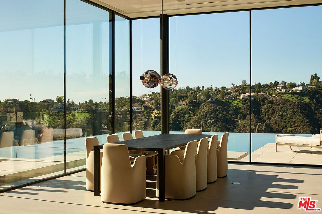 This interior shot showcases a modern dining room with floor-to-ceiling windows offering panoramic views of the landscape. A long, dark dining table is surrounded by light-colored chairs, and a unique pendant light fixture hangs above. The room's design emphasizes natural light and seamless integration with the outdoor environment, creating a luxurious and serene dining experience.