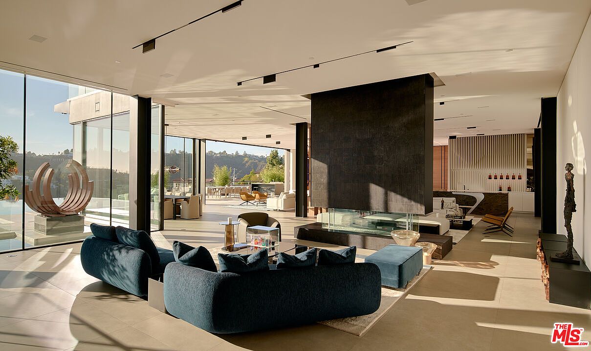 This is a spacious and modern living room featuring a large blue sectional sofa, a striking dark fireplace, and floor-to-ceiling windows that offer expansive views of the outdoors. The open floor plan seamlessly connects the living area to the outdoor patio, creating a bright and airy atmosphere. The room is decorated with contemporary art and minimalist furniture, enhancing the luxurious feel of the space.