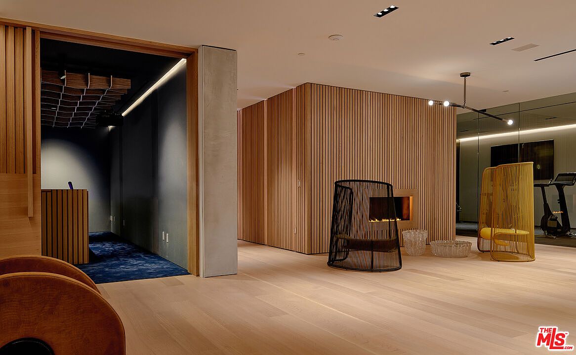 This interior shot showcases a modern home gym with a focus on natural wood elements and minimalist design. The space features wood-paneled walls, light hardwood flooring, and a mirrored wall reflecting exercise equipment. Stylish chairs and a fireplace add a touch of luxury to the workout area, creating an inviting and sophisticated atmosphere.