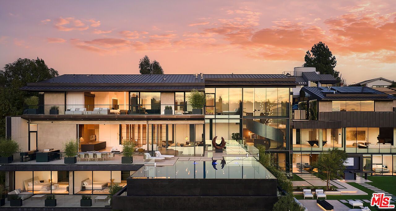 This is a stunning rear view of a modern luxury home at sunset, featuring multiple levels with floor-to-ceiling windows showcasing the interior spaces. A sleek infinity pool reflects the sky, and the outdoor area includes a patio with lounge seating and manicured landscaping, creating an inviting and sophisticated atmosphere.