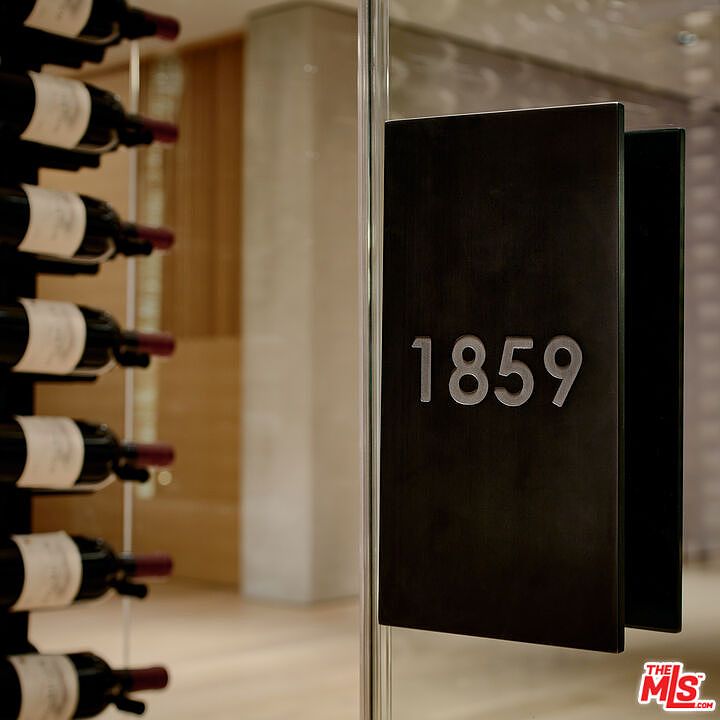 This image showcases a modern wine cellar, featuring a sleek glass door with a dark, rectangular handle displaying the number '1859'. Wine bottles are neatly arranged on a rack to the left, suggesting a sophisticated and organized storage space. The overall impression is one of luxury and attention to detail, highlighting a unique amenity for potential buyers.