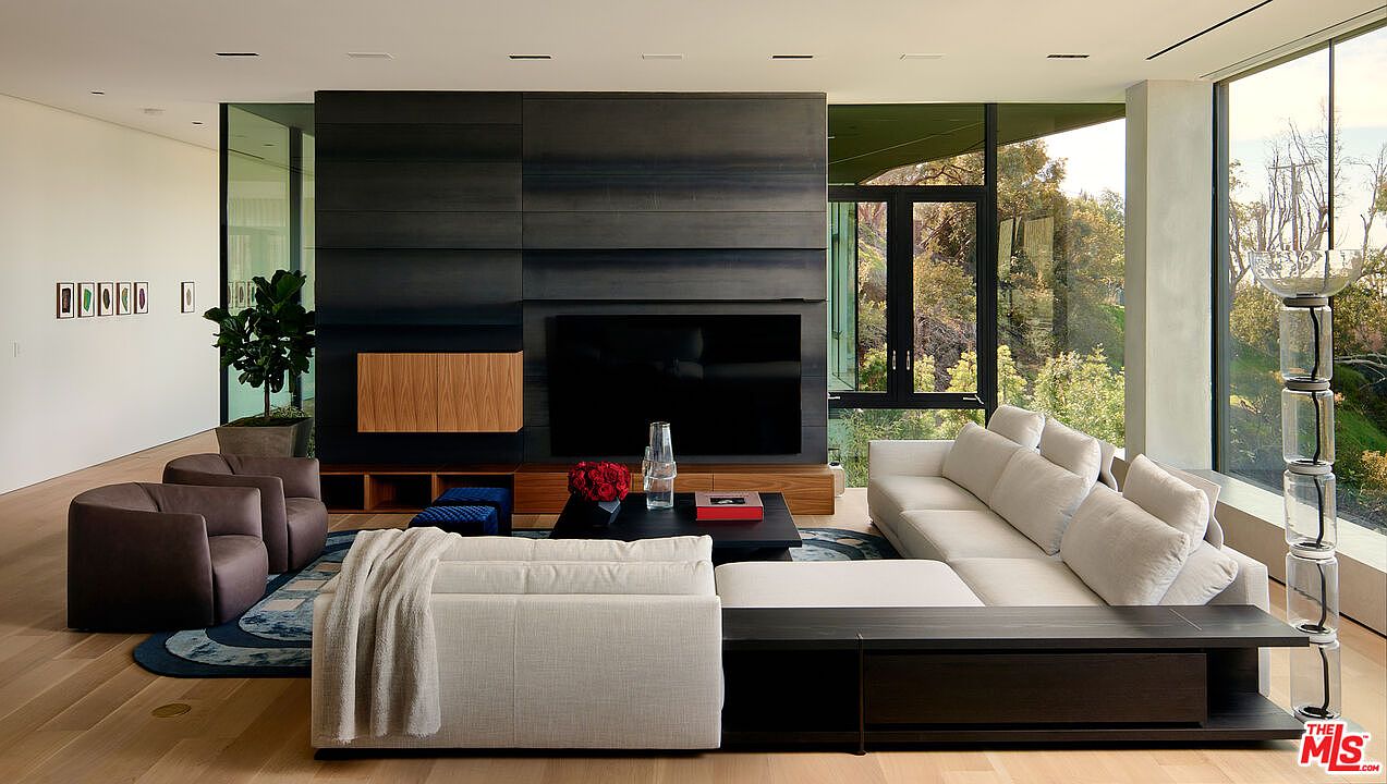 This is a modern living room featuring a large sectional sofa, two armchairs, and a dark-toned media center with a flat-screen TV. The room has floor-to-ceiling windows offering natural light and views of the surrounding greenery. The overall aesthetic is sleek and contemporary, emphasizing clean lines and a neutral color palette.
