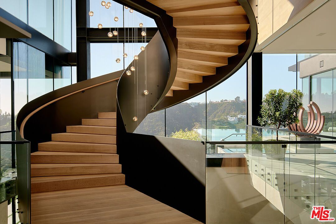This interior shot showcases a modern, curved staircase with wooden steps and a dark metal railing. A series of pendant lights hang elegantly above the stairs, adding a touch of sophistication. Large windows provide ample natural light and offer a glimpse of the outdoor scenery, enhancing the overall luxurious feel of the home.