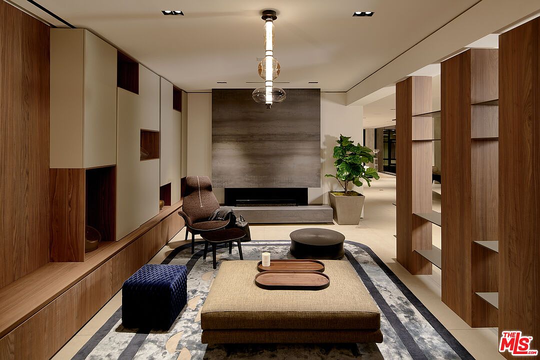 This is a modern living room featuring a neutral color palette with wood accents. The room includes a fireplace with a gray stone surround, a unique pendant light fixture, and a large area rug that defines the seating area. The space is furnished with a comfortable armchair, a large ottoman, and a blue upholstered cube, creating a cozy and stylish atmosphere.