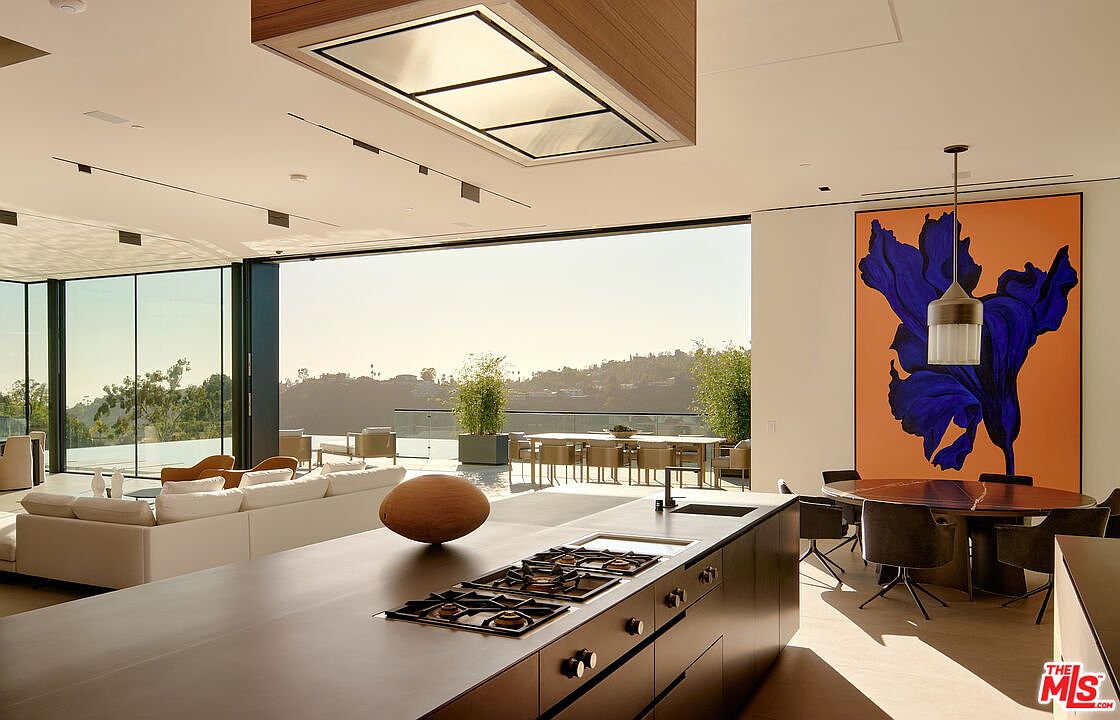 This interior shot showcases a modern, open-concept kitchen and living area with floor-to-ceiling windows offering expansive views. The kitchen features a sleek, dark-toned island with a built-in cooktop and a unique wooden decorative piece. A large, vibrant painting adds a pop of color to the dining area, which includes a round table and modern chairs, creating a luxurious and inviting atmosphere.