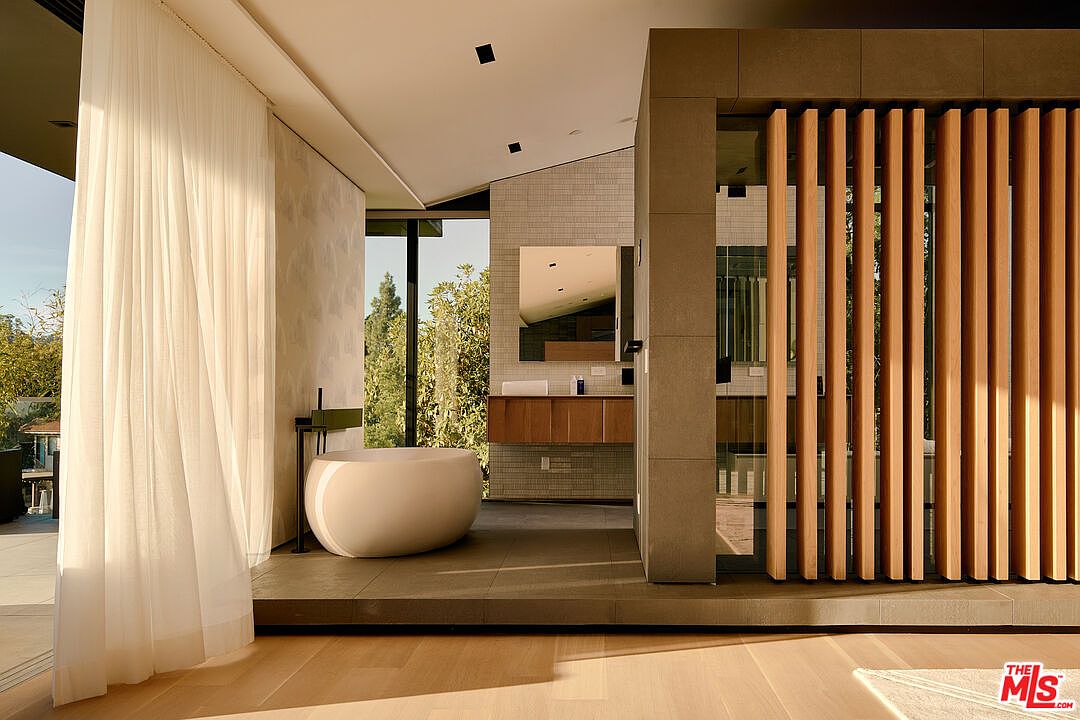 This is a luxurious primary bathroom featuring a freestanding oval bathtub next to a large window with sheer curtains. The bathroom has a modern design with a neutral color palette, including gray tile flooring and a wooden slat partition. A floating vanity with a mirror is visible in the background, contributing to the spa-like atmosphere.
