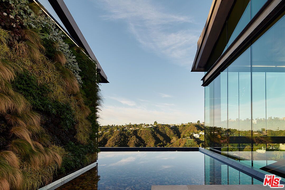 This image showcases a modern architectural design featuring a sleek infinity pool with clear water reflecting the sky and surrounding landscape. A vertical garden wall adds a touch of nature, contrasting with the clean lines of the glass-walled structure on the right. The overall impression is one of luxury, tranquility, and seamless integration with the environment.