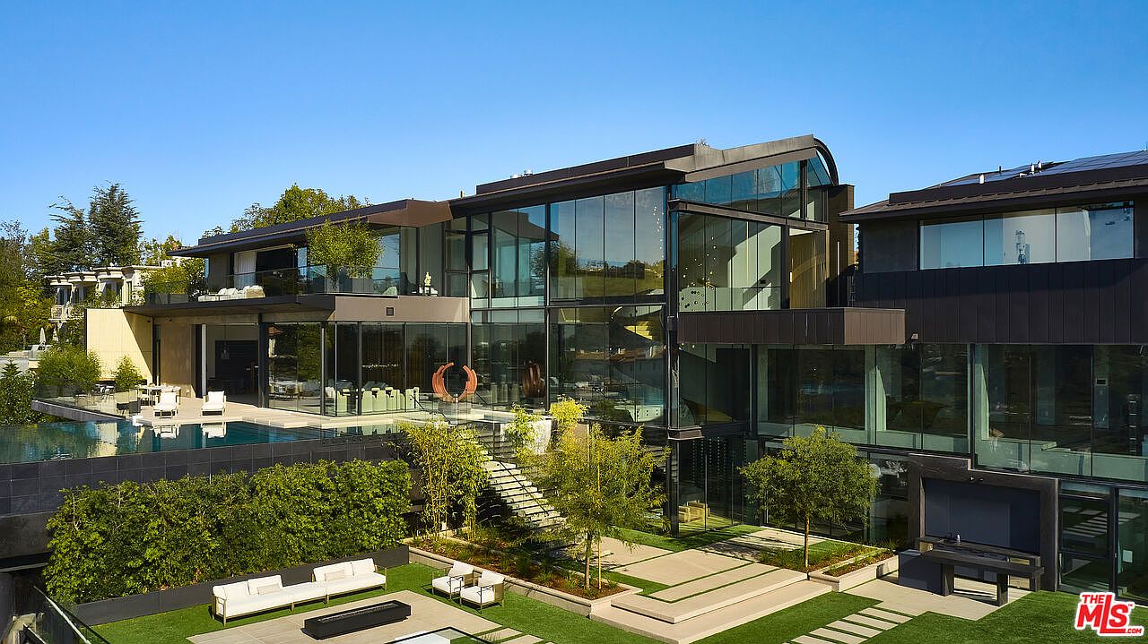 This is a rear view of a modern luxury home featuring extensive glass walls, a multi-level design, and a flat roof. The property includes a swimming pool, a meticulously landscaped yard with a patio area, and a staircase leading to an upper level. The architectural style emphasizes clean lines and a seamless integration of indoor and outdoor spaces.