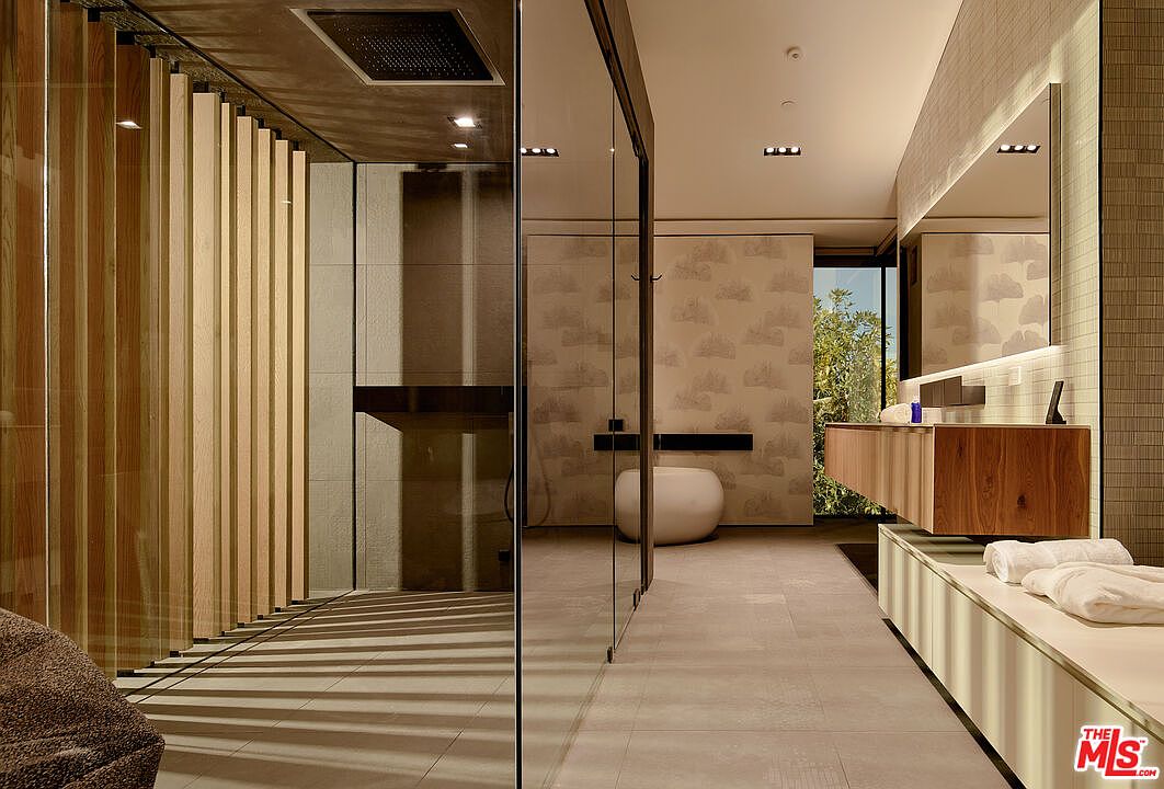 This is a modern primary bathroom featuring a walk-in shower with wooden slats, a floating vanity with a wood finish, and a large mirror. The bathroom has a neutral color palette with patterned wallpaper and tile accents. The space is well-lit and appears luxurious and spa-like.