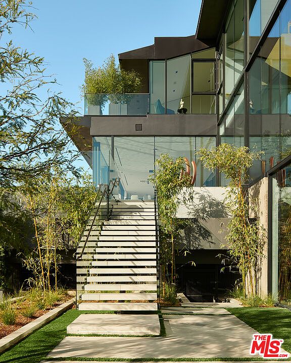 This is a striking front view of a modern home, showcasing a multi-level design with a prominent staircase leading to the main entrance. The exterior features a combination of glass, concrete, and dark metal accents, complemented by lush landscaping with bamboo and greenery. The architectural style is contemporary and sophisticated, creating a visually appealing and inviting facade.