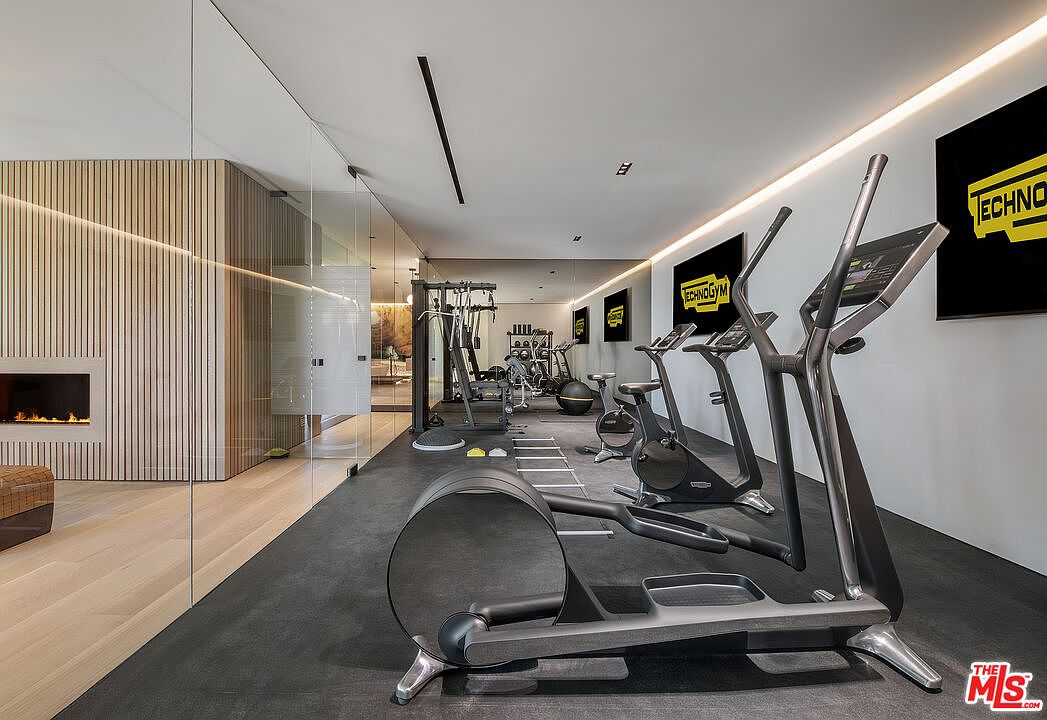 This is a modern home gym featuring state-of-the-art Technogym equipment, including elliptical machines and a weight training station. The room has a sleek design with a black rubber floor, a mirrored wall, and recessed lighting, creating a professional and motivating workout environment. A glass wall separates the gym from a lounge area with a fireplace, adding a touch of luxury.