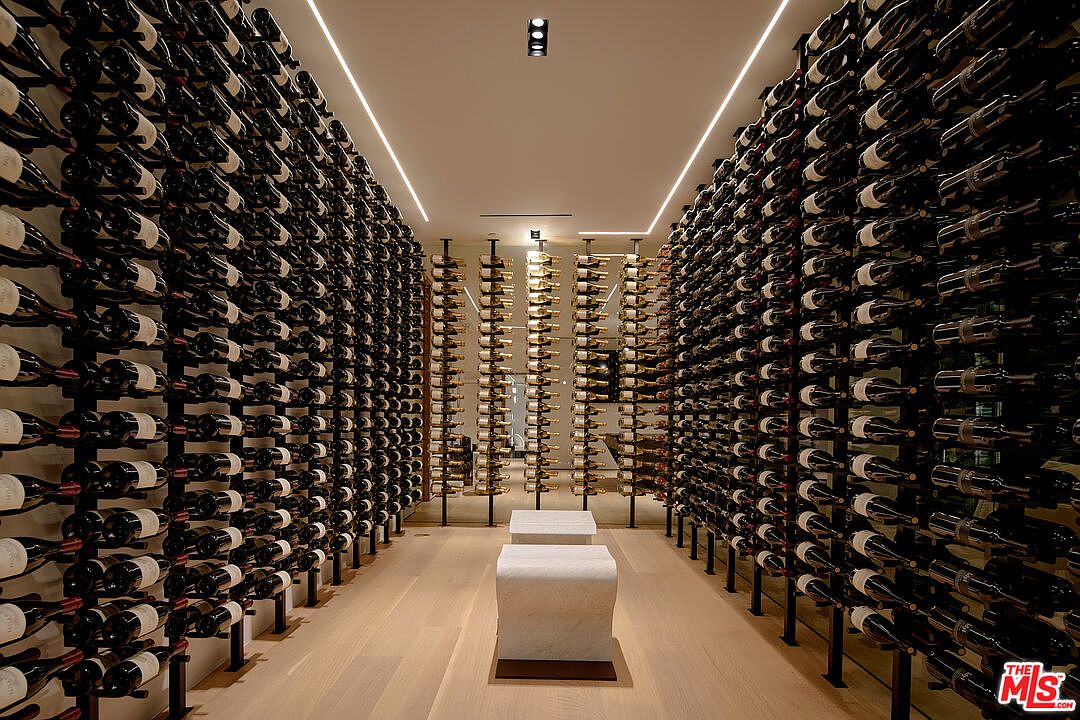 This is an interior shot of a well-stocked wine cellar. The cellar features rows upon rows of wine bottles on both sides, with a central aisle and minimalist seating. The lighting is modern, with linear fixtures, and the overall impression is one of luxury and sophistication.