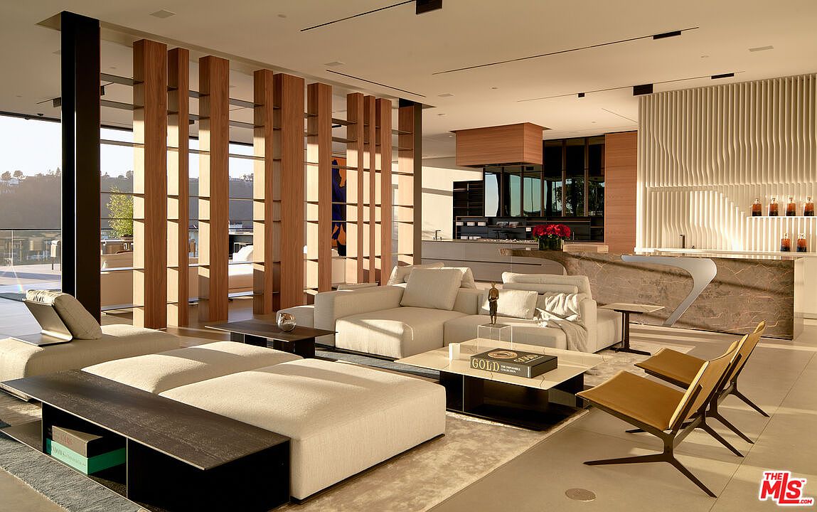 This is a spacious and modern living room featuring a large sectional sofa, a unique wooden slat wall, and floor-to-ceiling windows that provide ample natural light. The open-concept design seamlessly integrates the living area with the kitchen, creating a cohesive and inviting space. The neutral color palette and clean lines contribute to a sophisticated and contemporary aesthetic.