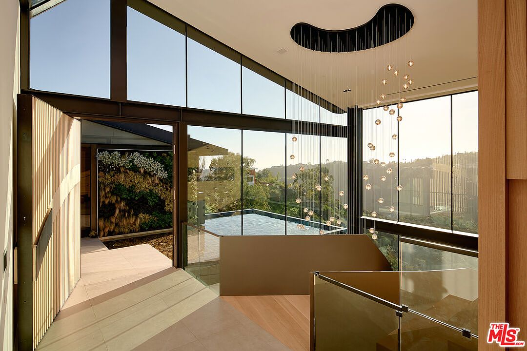 This interior shot showcases a modern hallway and staircase area with floor-to-ceiling windows offering views of a pool and lush greenery. The design features clean lines, glass railings, and a unique chandelier, creating a bright and airy atmosphere. The open doorway leads to an outdoor space with a vertical garden, enhancing the connection between indoor and outdoor living.