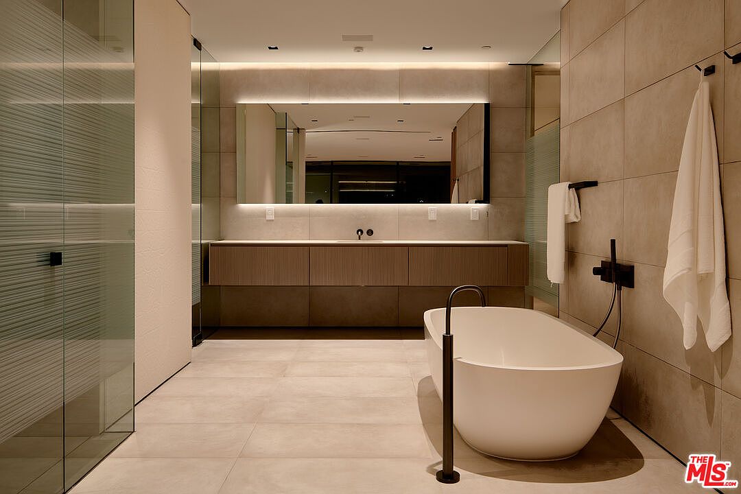 This is a modern primary bathroom featuring a freestanding white bathtub with a black floor-mounted faucet. The walls are covered in large format beige tiles, complemented by a floating vanity with a light wood finish and a large, illuminated mirror. A glass-enclosed shower is visible to the left, and a towel hangs on a black hook on the right wall, creating a spa-like atmosphere.