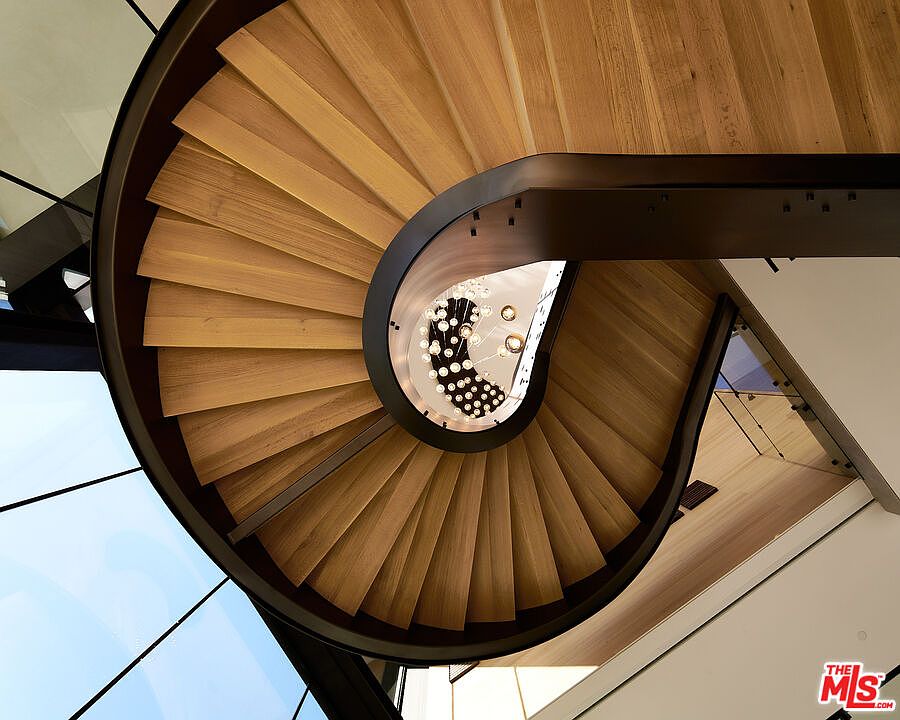 This image showcases a modern, spiral staircase viewed from below, emphasizing its architectural design. The wooden steps contrast with the dark metal framework, while a chandelier hangs in the center, adding a touch of elegance. The perspective highlights the staircase's unique shape and the interplay of light and shadow.