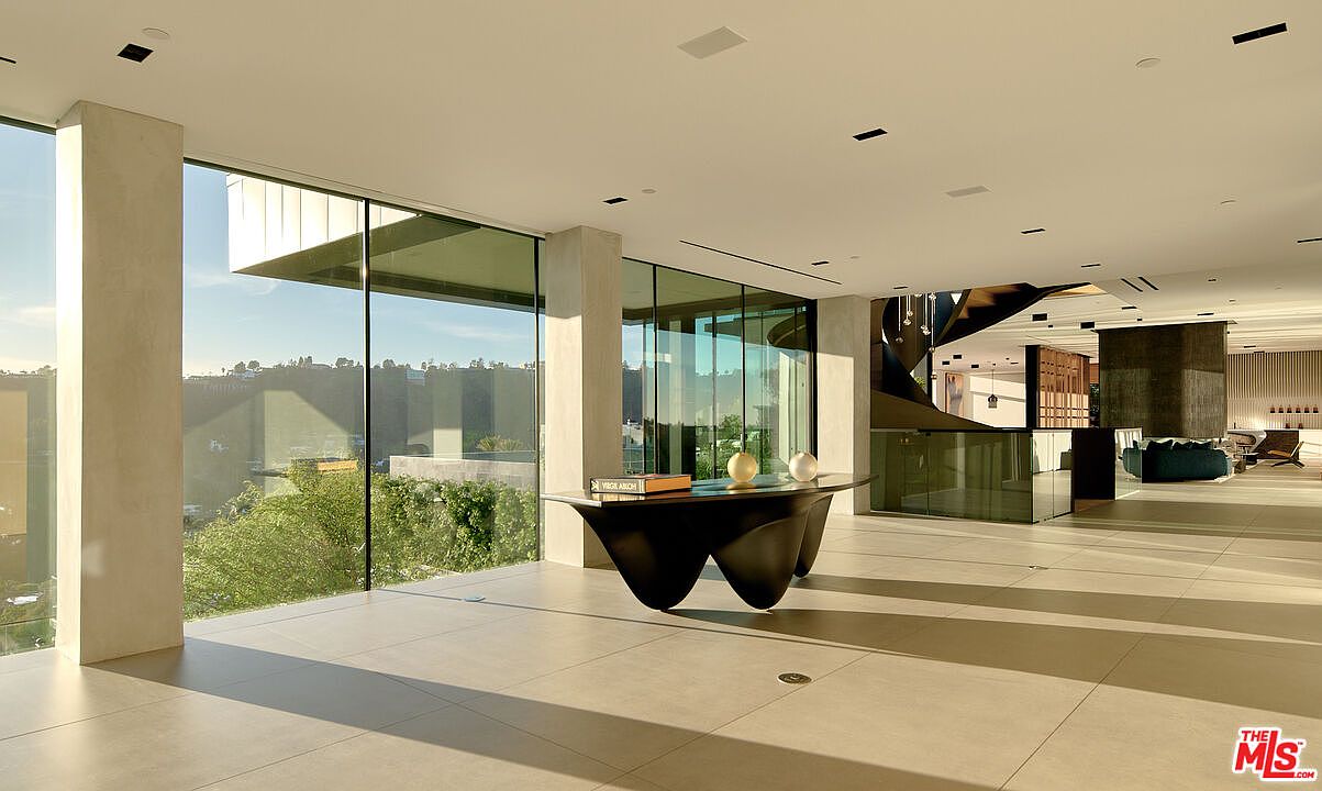 This interior shot showcases a modern hallway with large windows offering exterior views. A unique, dark-colored table with decorative objects sits prominently in the foreground, while a striking staircase design is visible in the background. The space is characterized by clean lines, neutral tones, and an abundance of natural light, creating an open and airy atmosphere.
