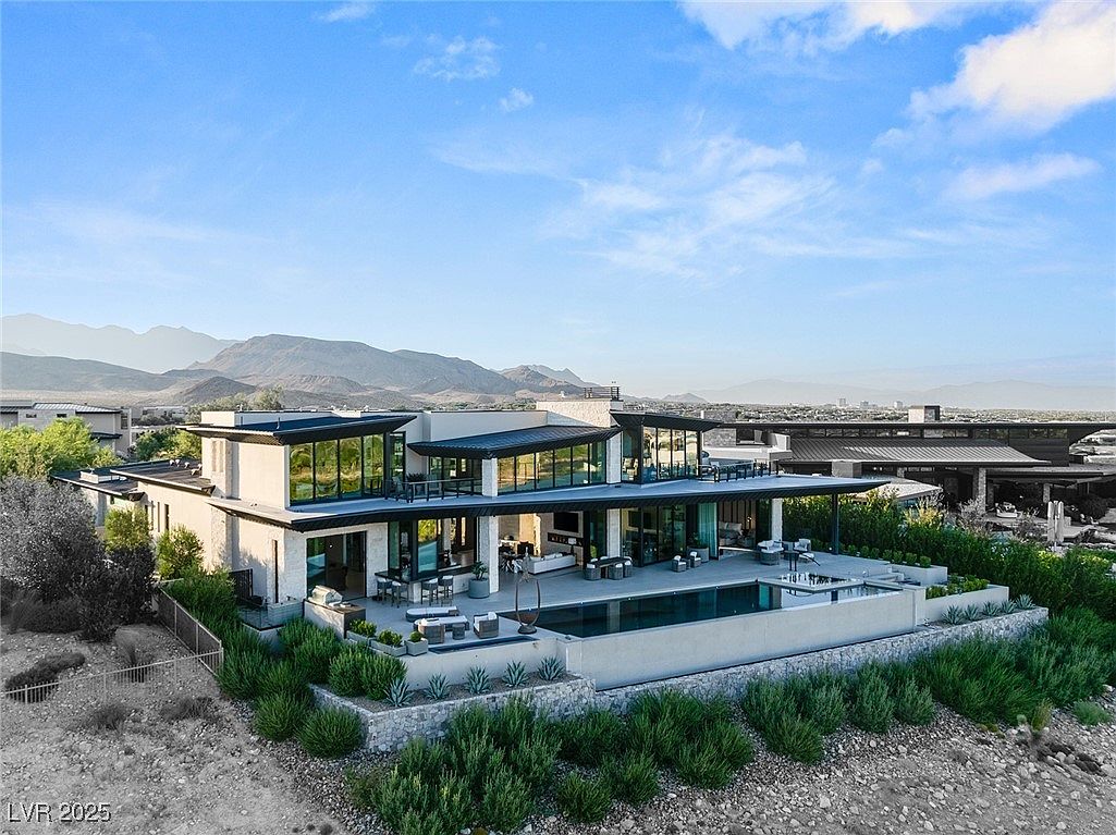 This aerial view showcases a luxurious modern home with a sleek, flat roof design and expansive windows. A stunning pool and patio area extend from the rear of the house, offering ample outdoor living space. The property is surrounded by desert landscaping and features a backdrop of mountains, creating a serene and private setting.