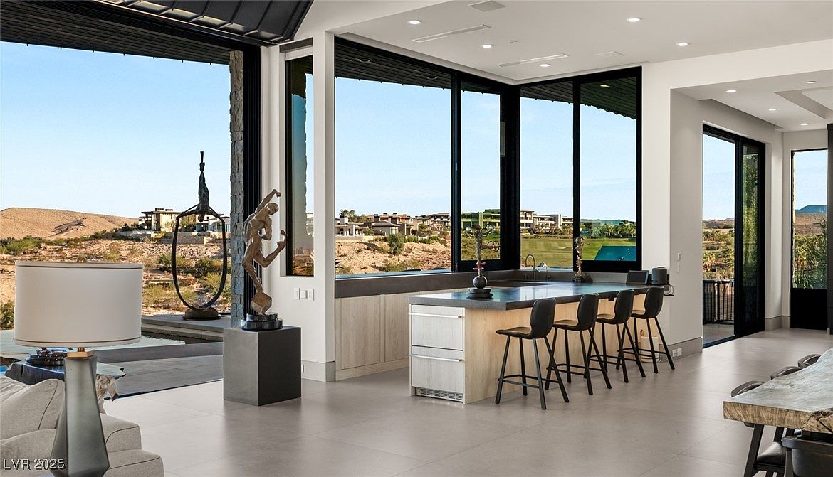 This interior shot showcases a modern kitchen with a large island and bar seating, framed by expansive windows offering panoramic views of the desert landscape. The kitchen features sleek cabinetry, a concrete countertop, and contemporary bar stools, creating a luxurious and inviting space. The open floor plan seamlessly connects the kitchen to the outdoor living area, enhancing the sense of spaciousness and natural light.