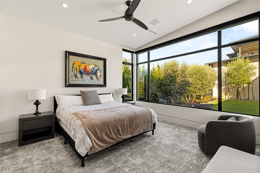 This is a primary bedroom featuring a large window with a view of the exterior greenery. The room is decorated with a modern aesthetic, including a ceiling fan, a painting of a rhino, and a gray patterned rug. The bed is neatly made with a neutral-toned blanket and decorative pillows, and there is a dark wood nightstand with a lamp on one side.