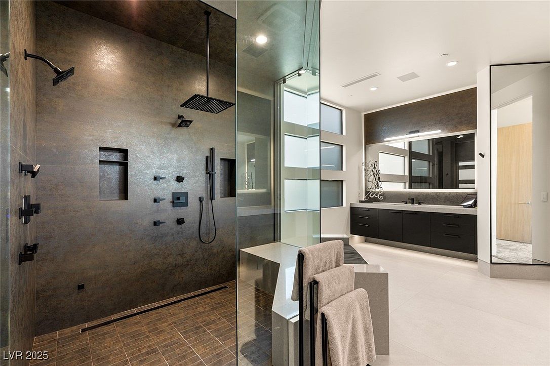 This is a luxurious primary bathroom featuring a spacious walk-in shower with multiple shower heads and a sleek, dark tile finish. The bathroom also includes a modern vanity with dark cabinetry, a large mirror, and contemporary fixtures. The overall design is sophisticated and spa-like, creating a relaxing and upscale atmosphere.