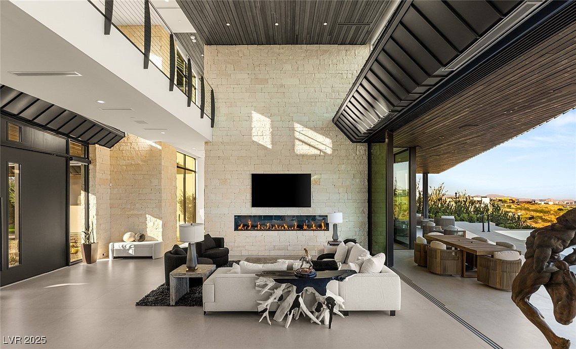 This is a spacious and luxurious living room featuring a modern fireplace with a large television mounted above it. The room has high ceilings, stone accent walls, and large windows that provide ample natural light. The furniture is contemporary and stylish, creating an inviting and sophisticated atmosphere.