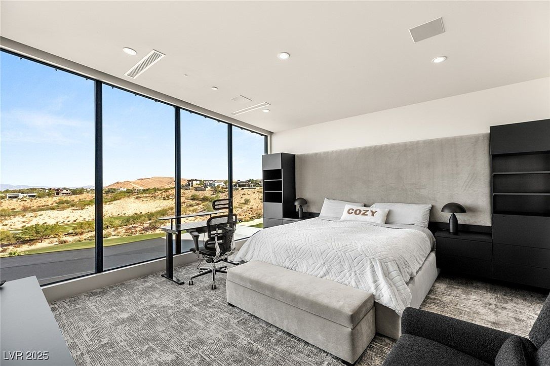 This is a primary bedroom featuring a large window with a view of the landscape. The room includes a bed with a headboard, a desk with a chair, and built-in shelving units. The color palette is neutral, with gray and black tones, creating a modern and sophisticated atmosphere.