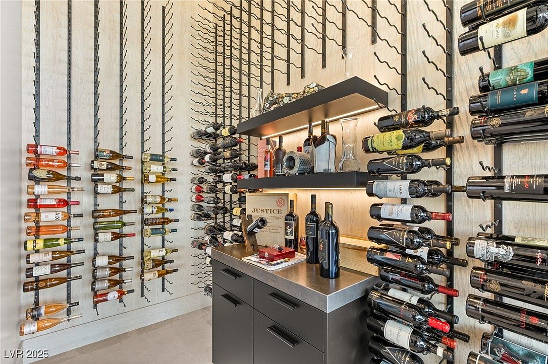 This is a well-organized wine cellar featuring a modern design with a focus on wine storage and display. The cellar includes multiple wine racks, a countertop with storage cabinets, and shelving with decorative items. The lighting highlights the wine collection and creates an inviting atmosphere, perfect for showcasing a curated selection.