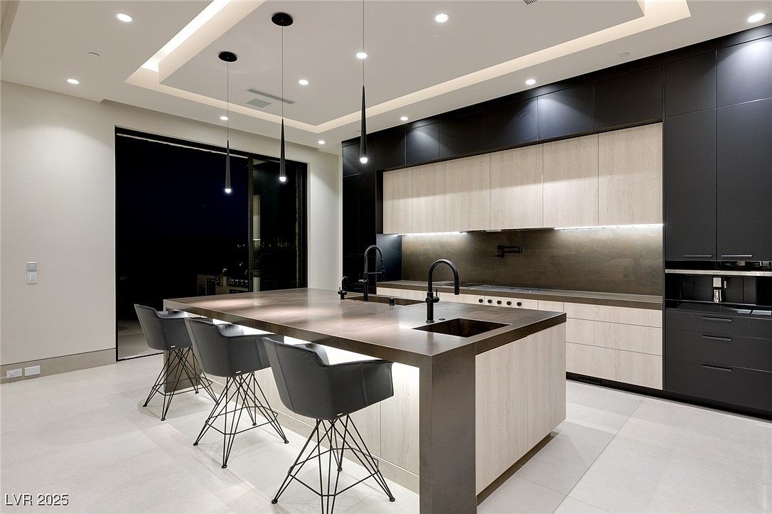 This is a modern kitchen featuring a large island with seating, sleek black and light wood cabinetry, and contemporary pendant lighting. The kitchen is well-lit with recessed lighting and features high-end appliances. A large sliding glass door provides access to the outside, creating an open and airy feel.