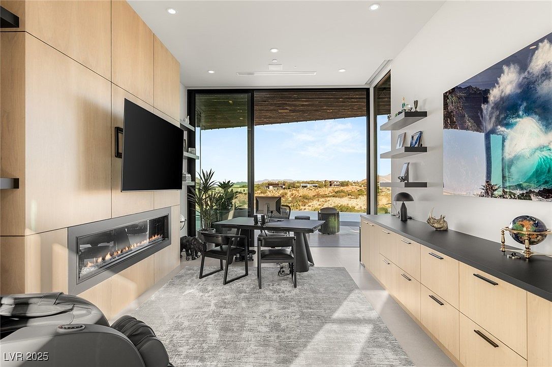 This is a modern office space featuring a linear fireplace, a wall-mounted television, and a large window offering an expansive view of the landscape. The room includes a sleek desk with chairs, built-in cabinetry, and a contemporary art piece, creating a sophisticated and functional workspace. The color palette is neutral with wood accents, contributing to a calm and professional atmosphere.