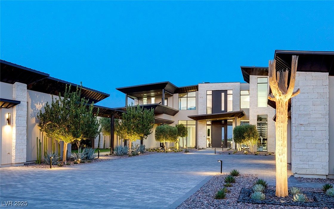 This is a front exterior view of a modern luxury home. The property features a large paved driveway, manicured landscaping with trees and succulents, and a striking architectural design with clean lines and a combination of stone and stucco finishes. The home's facade includes large windows and a covered entryway, creating an inviting and sophisticated curb appeal.