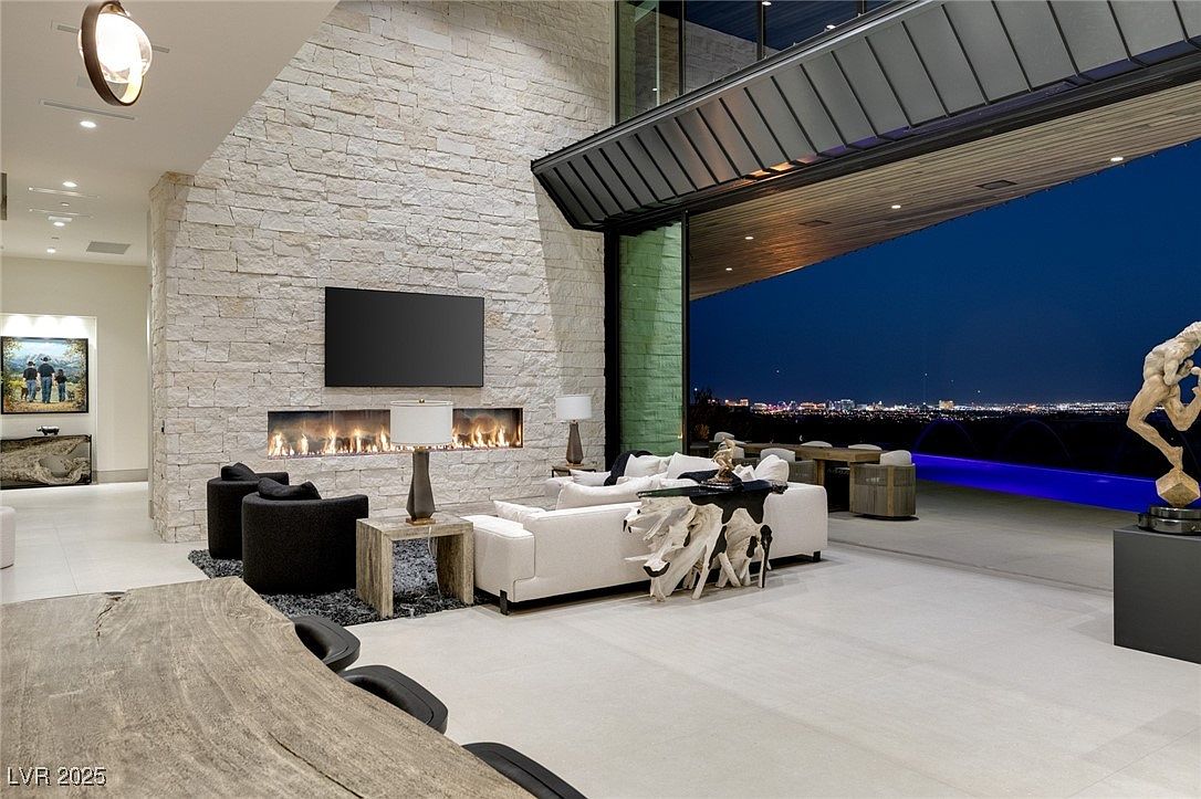 This is a luxurious living room featuring a stone accent wall with a modern fireplace and a large flat-screen TV. The room opens to an outdoor patio with a pool and city views, creating a seamless indoor-outdoor living experience. The decor includes contemporary furniture and artistic elements, contributing to an elegant and sophisticated ambiance.