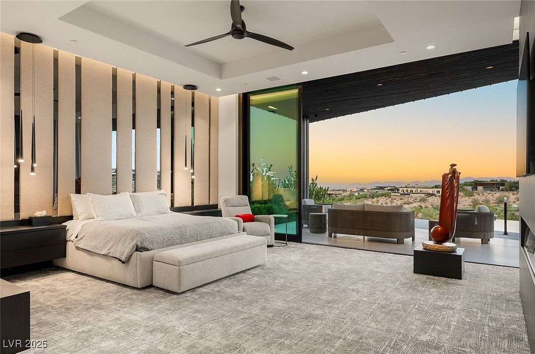 This is a luxurious primary bedroom featuring a large bed with a padded headboard and a bench at the foot. The room has a modern design with a neutral color palette, accented by dark wood elements and sleek lighting fixtures. A large sliding glass door opens to an outdoor patio area with seating, offering stunning views of the landscape and sunset.