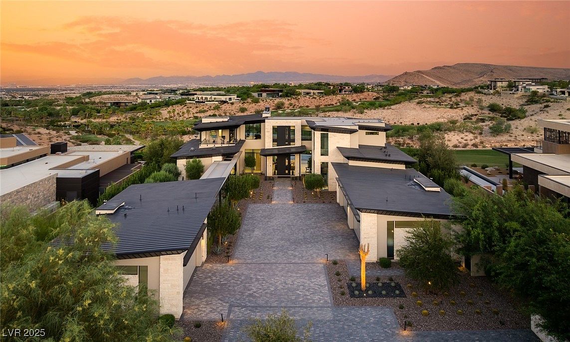 This aerial view showcases a luxurious modern home with a striking architectural design, featuring a combination of flat and angled roofs, light-colored stone or stucco siding, and expansive windows. The property includes a spacious driveway paved with interlocking stones, meticulously landscaped grounds, and is situated in a scenic location with views of distant mountains and desert landscape, creating an impression of exclusivity and high-end living.