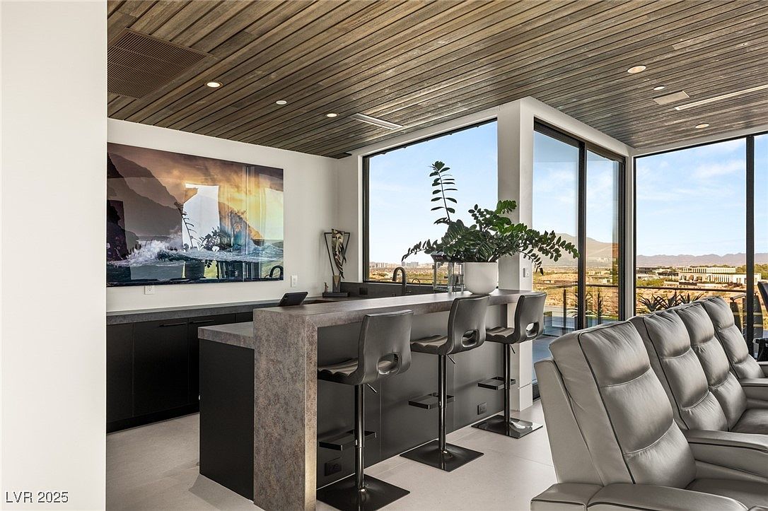 This interior shot showcases a modern living room with a bar area, featuring sleek bar stools and a countertop with a sink. Large windows offer panoramic views of the landscape, while a comfortable gray leather sofa provides a relaxing seating area. The room is well-lit with recessed lighting and natural light, creating a sophisticated and inviting atmosphere.