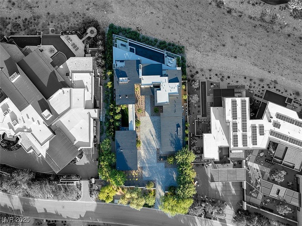 This aerial shot showcases a modern luxury home with a dark roof, light-colored exterior walls, and a unique architectural design. The property features a swimming pool, well-manicured landscaping, and a spacious driveway. The surrounding area includes other high-end homes and desert terrain, emphasizing the property's exclusive location.