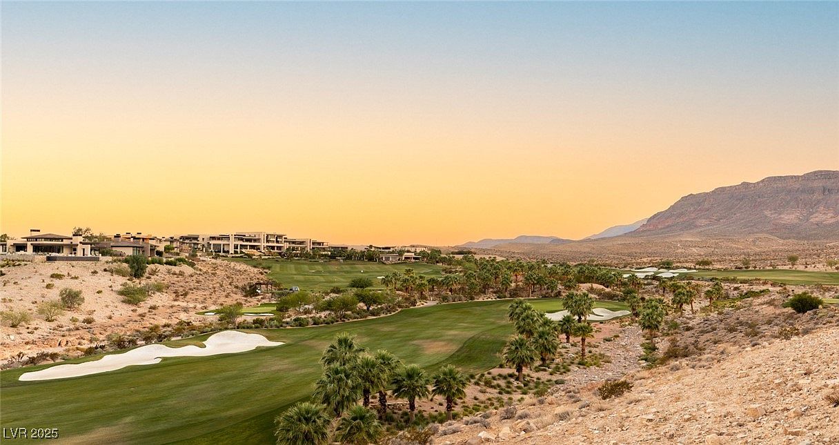 This image showcases a luxurious golf course community at sunset, featuring meticulously manicured green fairways, palm trees, and sand traps. Elegant homes are nestled on the hillside overlooking the course, with a backdrop of desert mountains under a vibrant sky. The scene evokes a sense of exclusivity and upscale living.