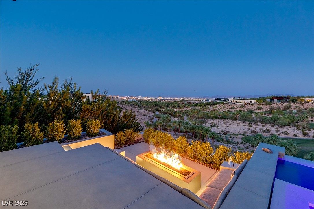 This image showcases a luxurious outdoor patio area at dusk, featuring a modern fire pit as a focal point. The patio is surrounded by manicured greenery and offers panoramic views of the city skyline in the distance. The presence of a pool and comfortable seating suggests an ideal space for relaxation and entertainment.