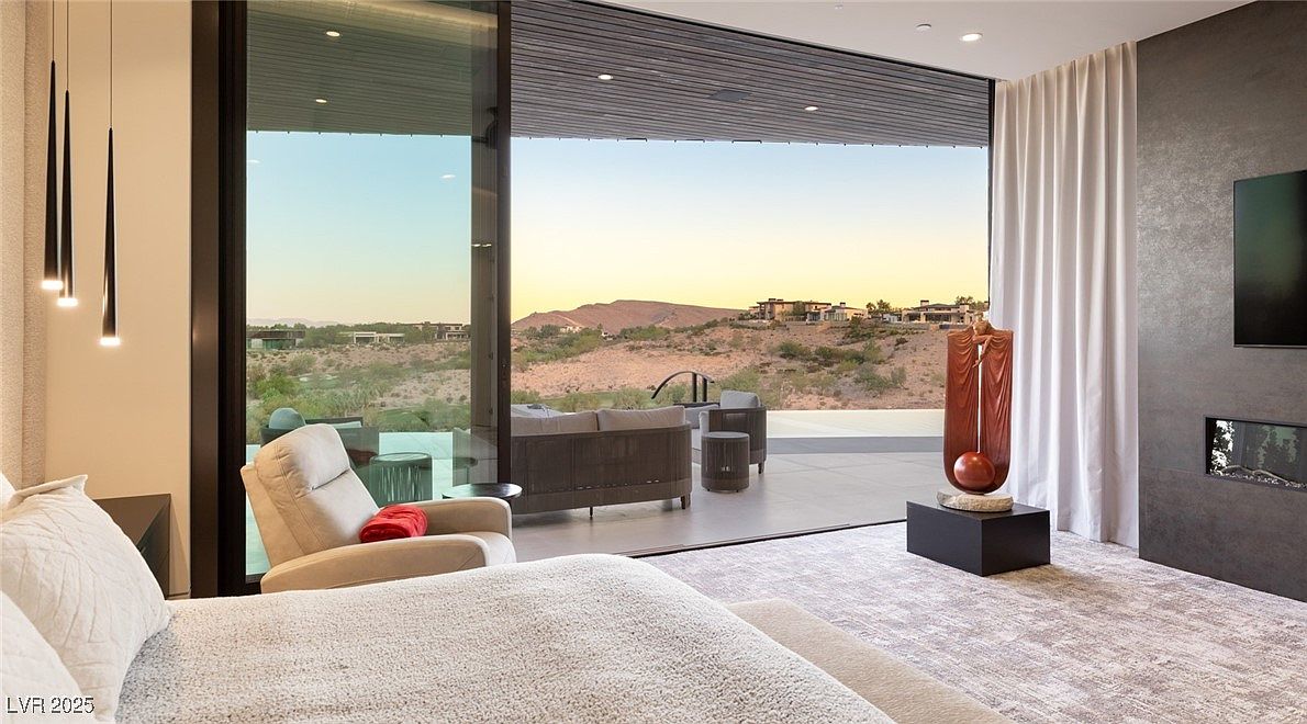 This is a luxurious primary bedroom featuring a large bed with plush bedding, a modern fireplace, and a striking sculpture. Floor-to-ceiling windows offer panoramic views of the desert landscape, seamlessly blending indoor and outdoor living. The room is decorated in neutral tones, creating a serene and sophisticated atmosphere.