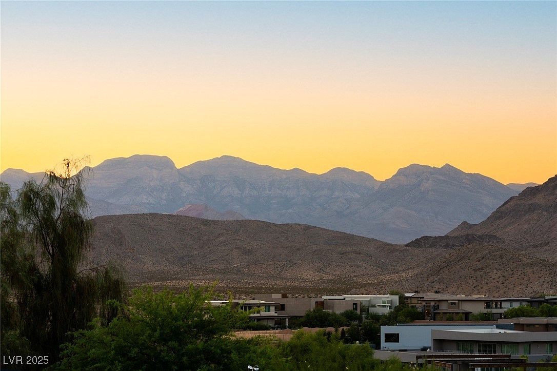 This image showcases a modern home nestled in a desert landscape, with a stunning mountain range as a backdrop. The architecture features clean lines and a neutral color palette, blending seamlessly with the natural environment. Lush greenery surrounds the property, adding a touch of vibrancy to the scene, while the golden hour lighting enhances the overall warmth and appeal.