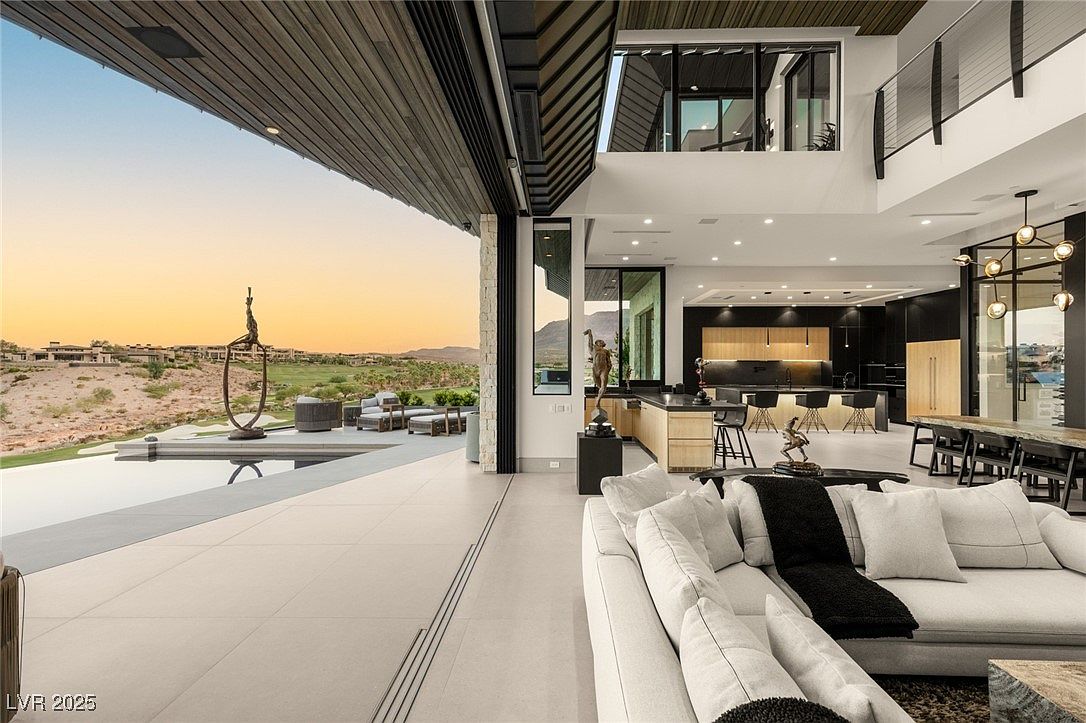 This image showcases a luxurious living room that seamlessly blends indoor and outdoor spaces. The open floor plan features a modern kitchen in the background, a dining area, and a comfortable living area with a large sectional sofa. The expansive sliding glass doors open to a patio with a pool and stunning views, creating an inviting and sophisticated atmosphere.